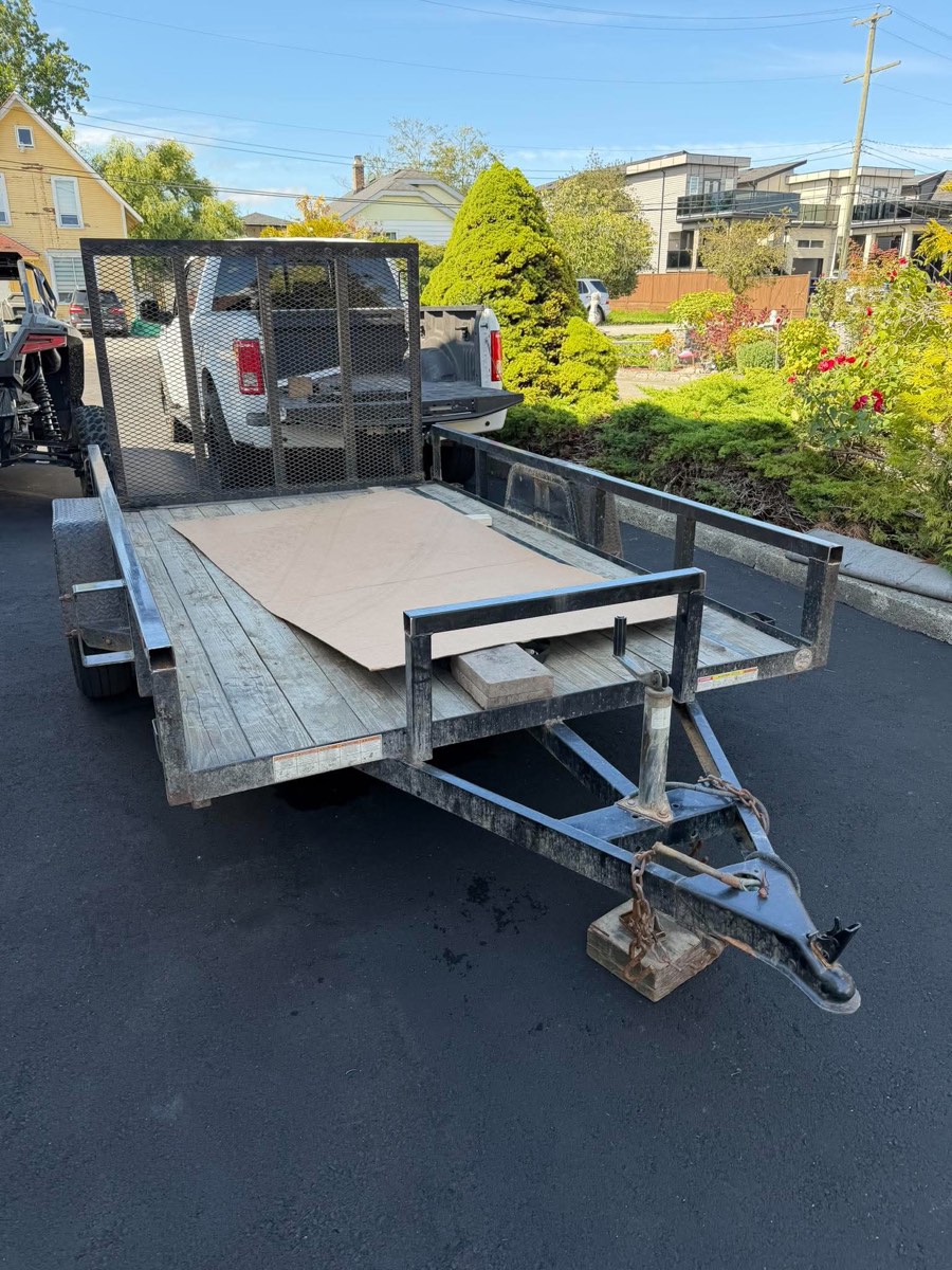 Standard wood-deck utility trailer unhitched in a driveway, side-by-side parked beside it